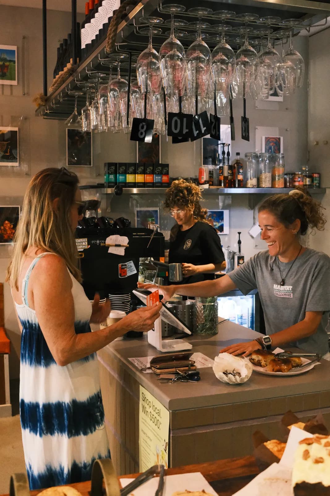 Customers at the counter, served by the team, under the hanging wine glasses.